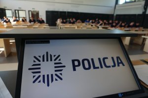 ekran z napisem policja a w tle policjanci siedzący przy stolikach na auli