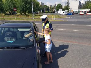 policjantka z dziewczynką wręczają siedzącemu za kierownicą mężczyźnie laurkę