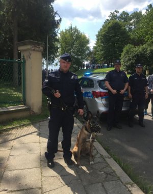 Policjant umundurowany-przewodnik z psem służbowym, w tle część radiowozu policyjnego oraz dwóch umundurowanych funkcjonariuszy