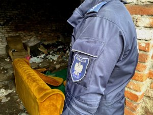 ramie umundurowanego policjanta, w tle pustostan pełen śmieci