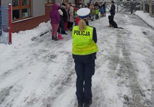 umundurowana policjantka w kamizelce odblaskowej stoi tyłem do fotografującego i patrzy w kierunku grupy osób i przewodnika psa służbowego