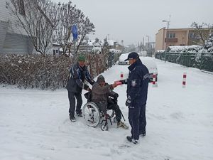 Umundurowany policjant na zaśnieżonym  chodniku przekazuje apteczkę osobie na wózku inwalidzkim prowadzonym przez drugiego mężczyznę