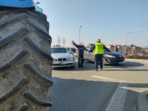 Koło od ciągnika, w tle policjant rozmawiający z obywatelem, który wysiadł z samochodu i podnosi ręce do góry