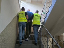 zdjęcie kolorowe policjanci w odblaskowych kamizelkach z napisem policja prowadzą po schodach zatrzymanego mężczyznę